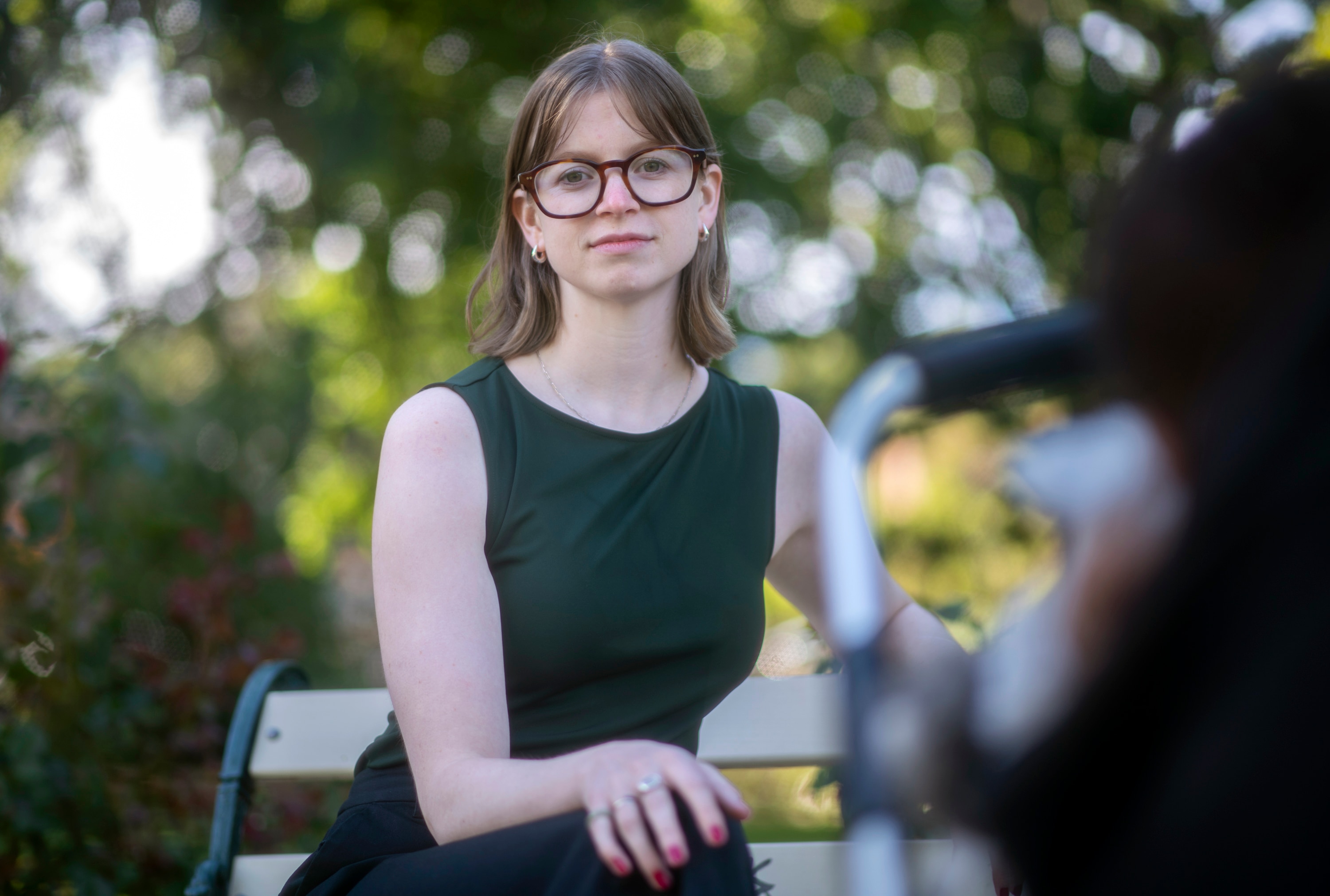 Jarin White, with glasses and a green dress, sits on a park bench with soft greenery in the background.