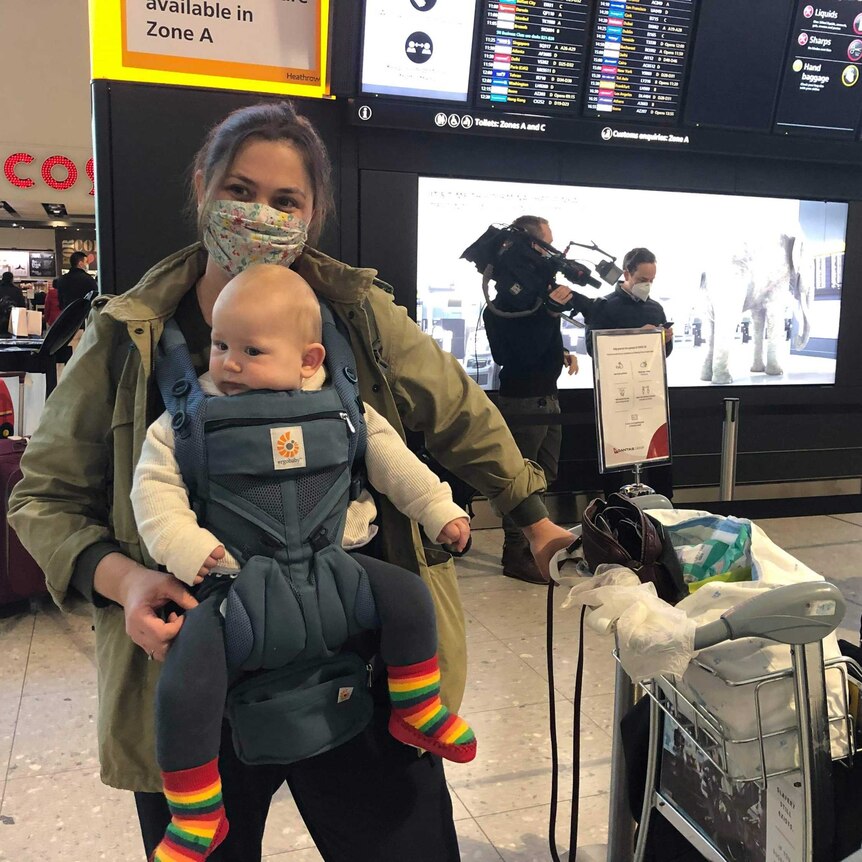 A mother and her baby at an airport look to the camera. The mother is wearing a mask.