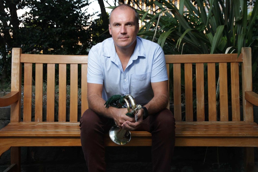 A man wearing a light blue shirt and maroon trousers sits on a park bench holding a bugle.