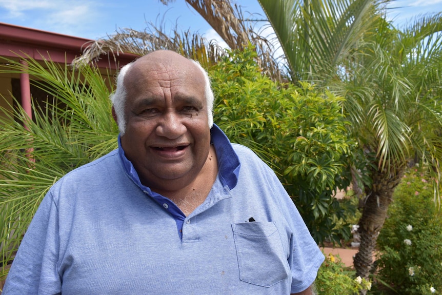 A tall man with dark skin and white hair standing in a garden with green trees behind him