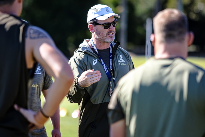 A coach wearing sunglasses and a hat gesticulates to players