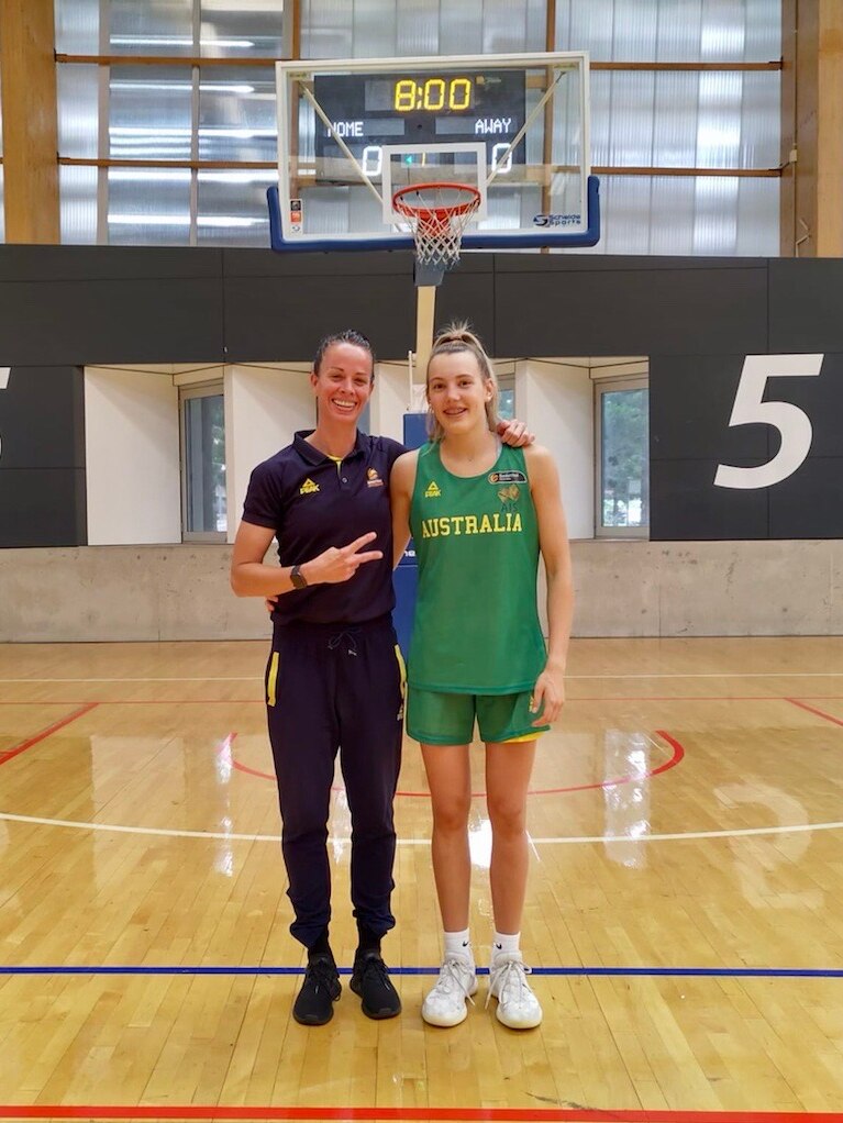 A teenager, wearing green and gold, standing with a woman on a basketball court.
