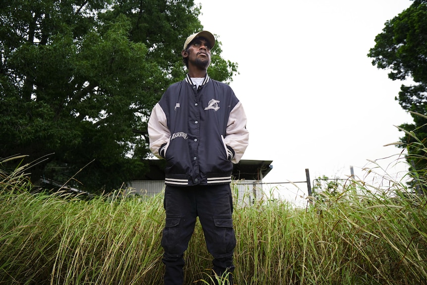 Man standing in tall grass, staring into distance, he's wearing a cap and jacket.