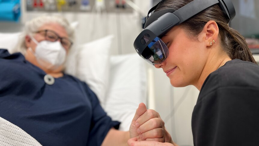 Heidi wears a sleek black VR headset as she holds the hand of an elderly patient lying in a hospital bed.