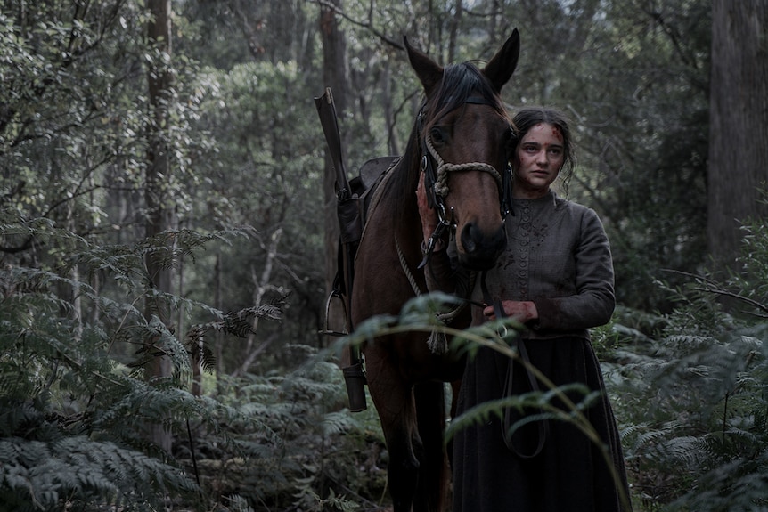 A woman with worried expression and covered in some blood stands closely with horse in Australian bushlands.