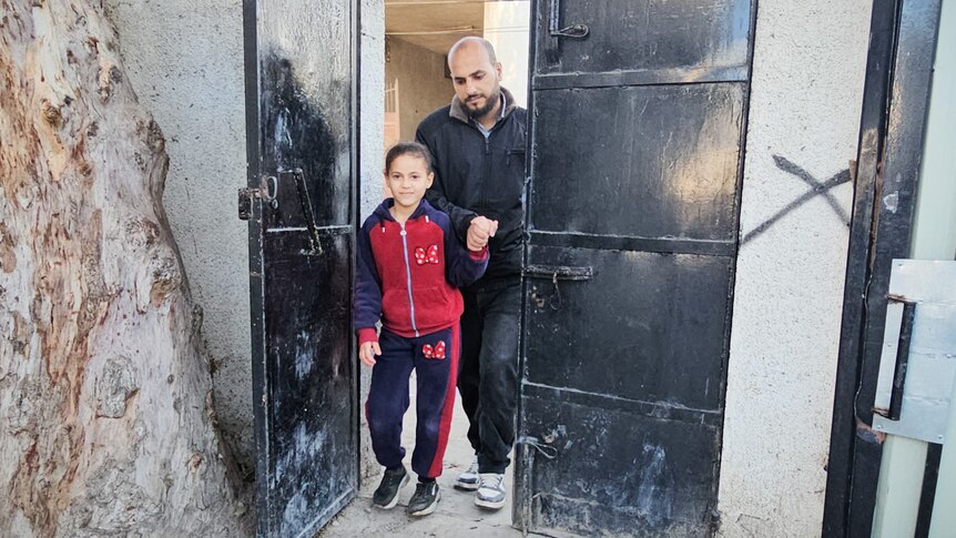 A young girl wearing a red jumper holds her father's hand while walking through a door.