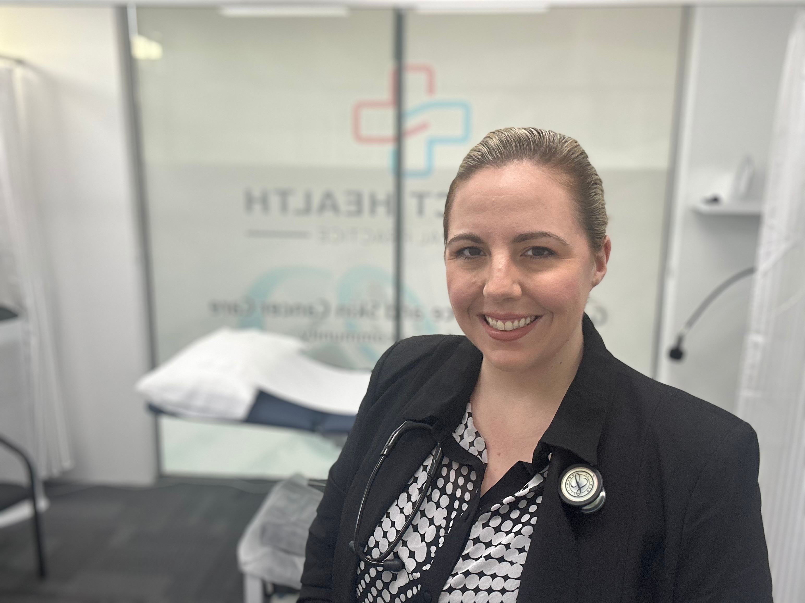A smiling woman standing in front of a medical bed with a stethoscope around her neck
