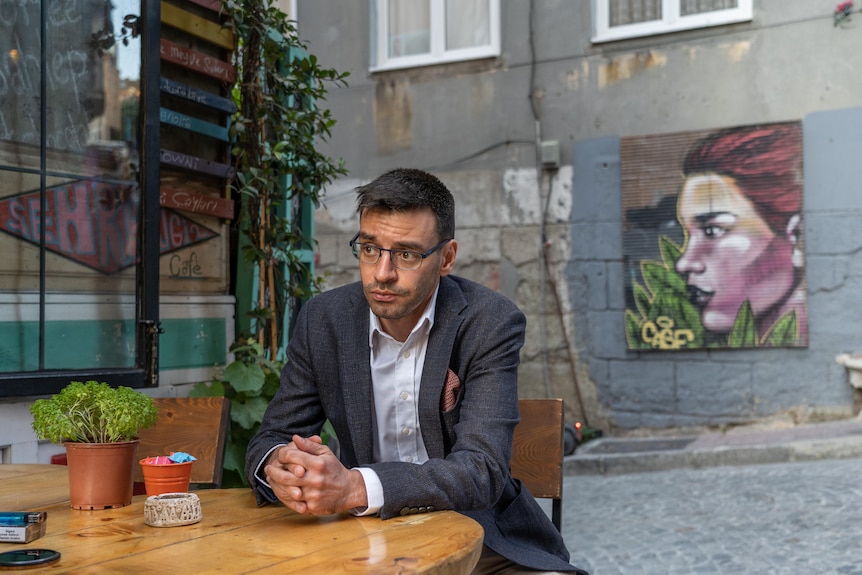 A man wearing a smart blazer and glasses sits at an outdoor cafe table in an industrial alley
