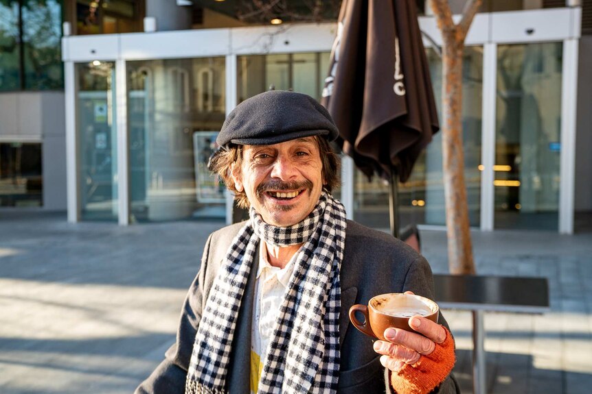 A man in a scarf and flat cap is smiling with a coffee in his hand.
