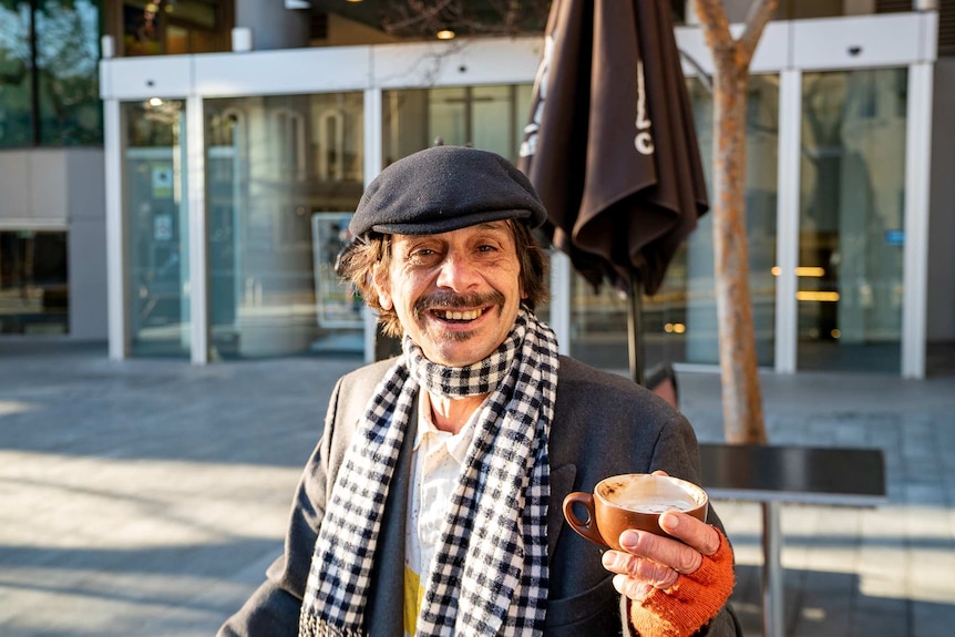A man in a scarf and flat cap is smiling with a coffee in his hand.