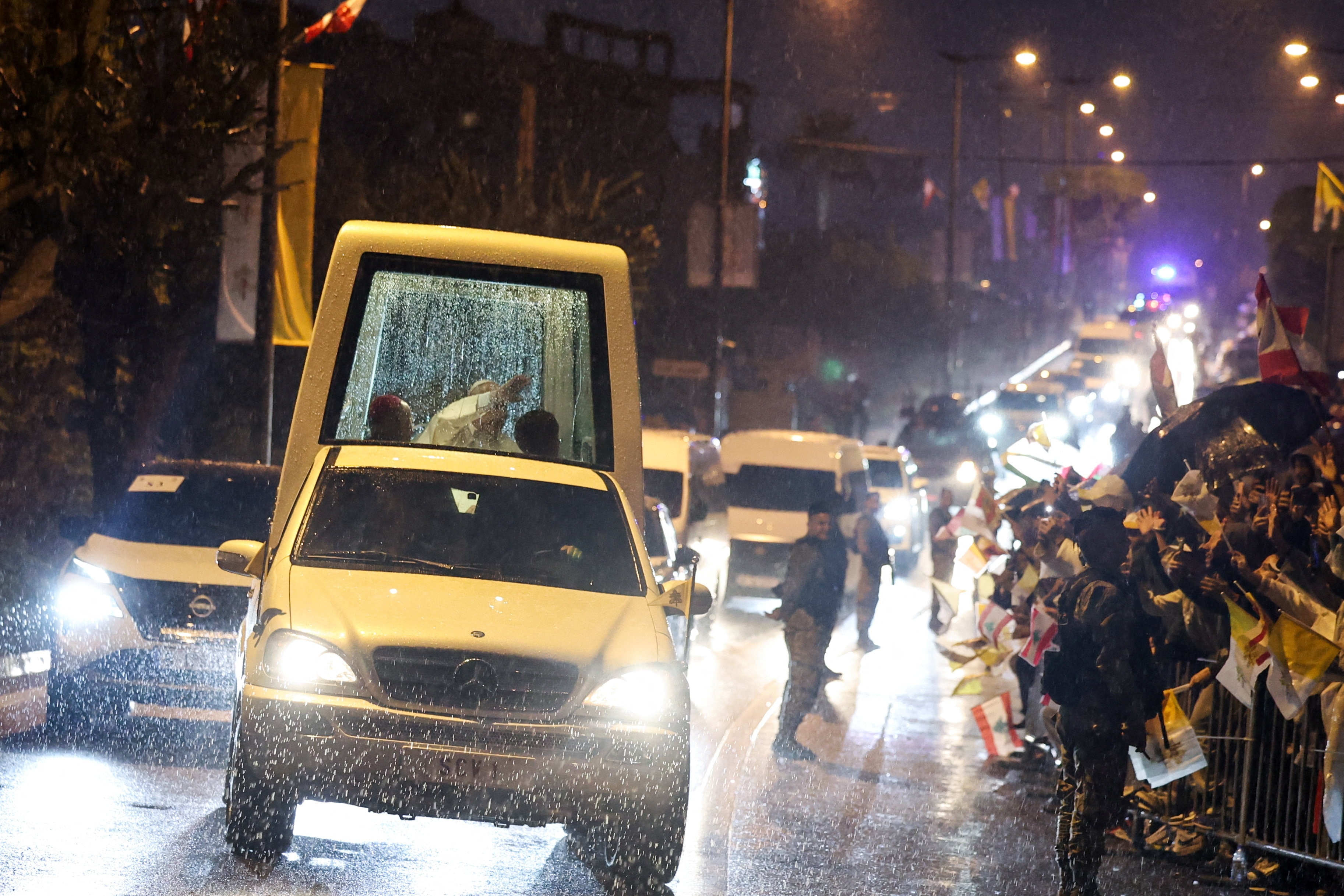 The pope sitting in the glass box of the popemobile waving at crowds of people behind fences along the road in the rain