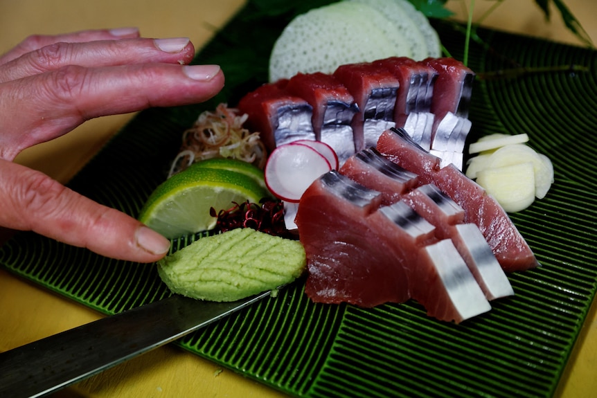 A person's hand holds bits of wasabi next to cut up pieces of katsuo on a mat at a restaurant