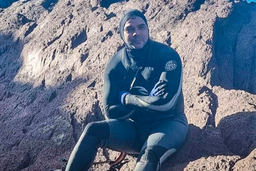 Man in wetsuit and hood sitting on rocks with arms folded, smiling.