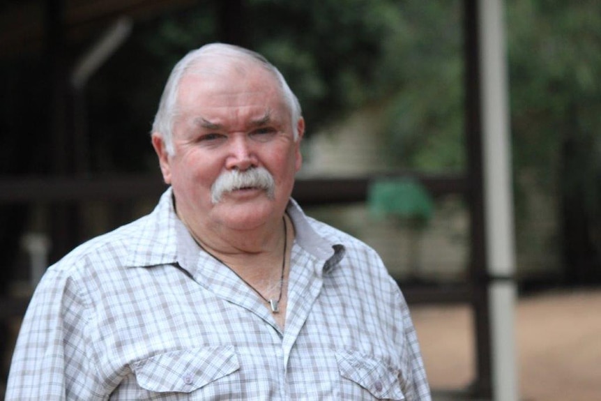 An older man with a grey moustache stands in front of the camera.