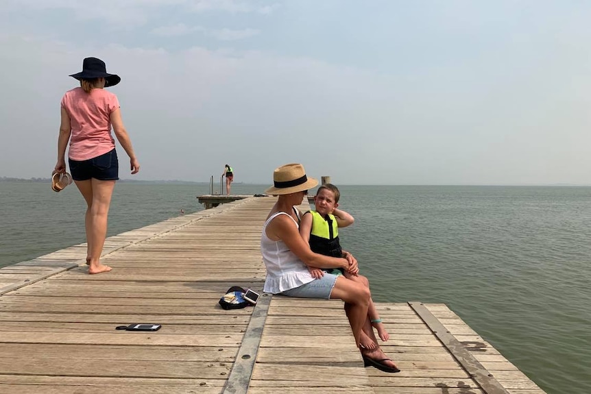 A boy is sitting on his mum's lap on a jetty. There is a lady walking behind them and water all around them.