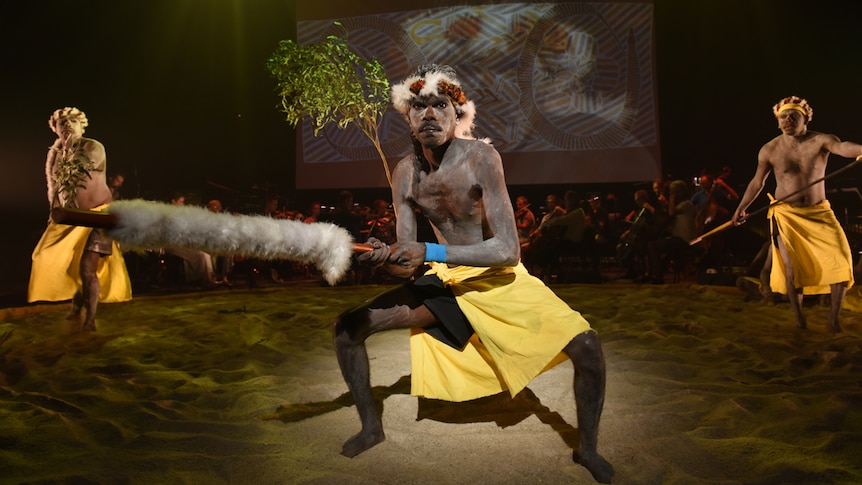 Three men with ochre on faces and torsos dancing on sand circle on stage with projection of Yolngu artwork behind them and tree.