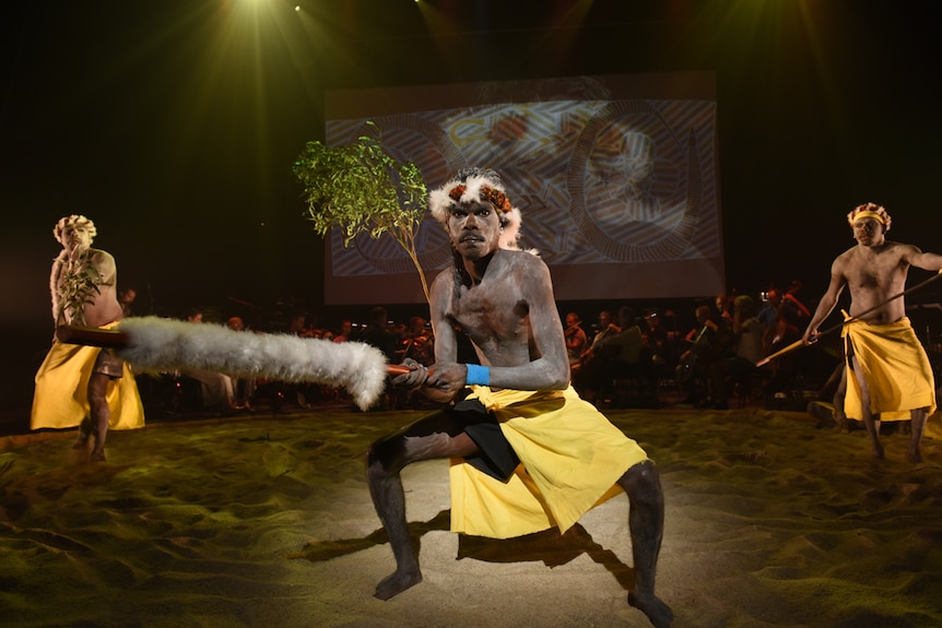 Three men with ochre on faces and torsos dancing on sand circle on stage with projection of Yolngu artwork behind them and tree.