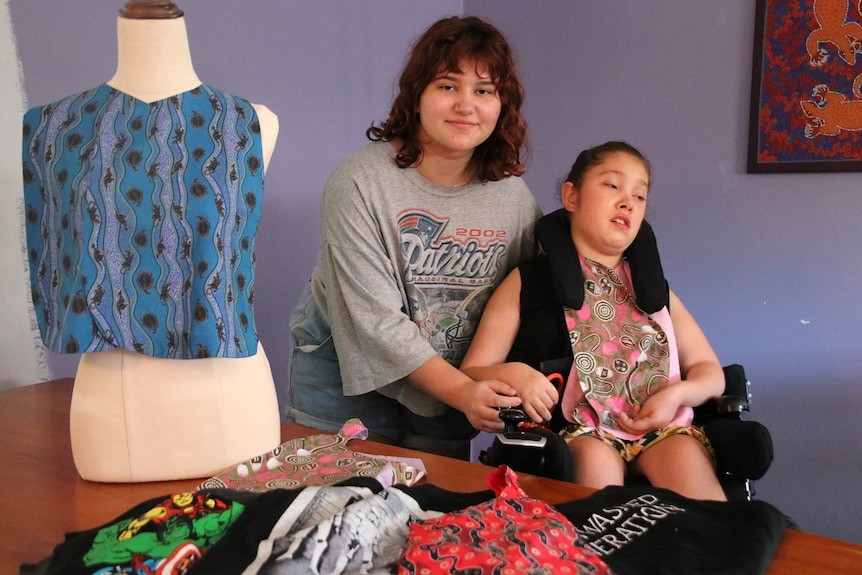 A young girl stands next to a girl in a wheelchair with clothing fabrics draped on a manniken.