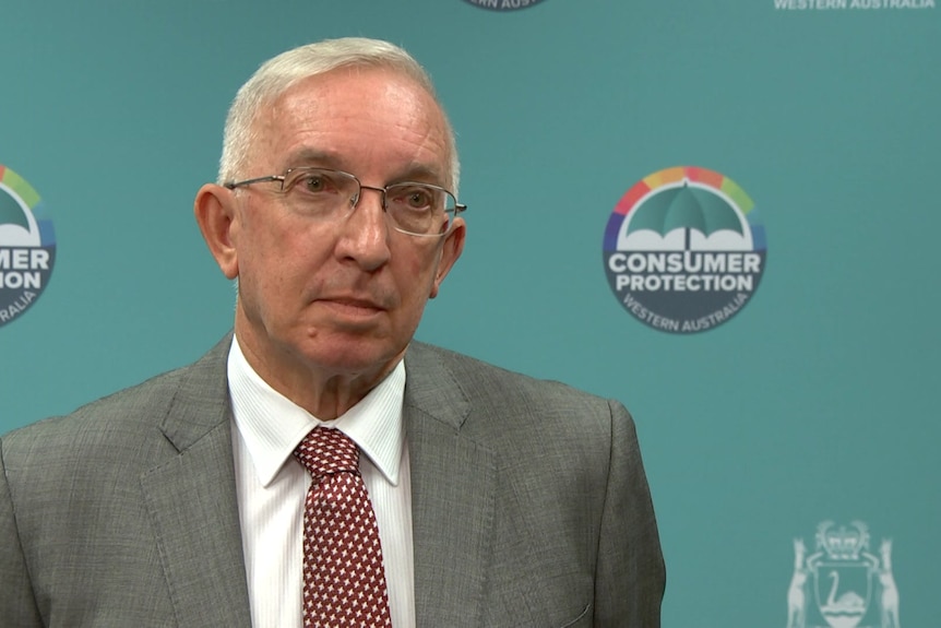 A man with grey hair and a red tie speaking to the media in front of a green background.