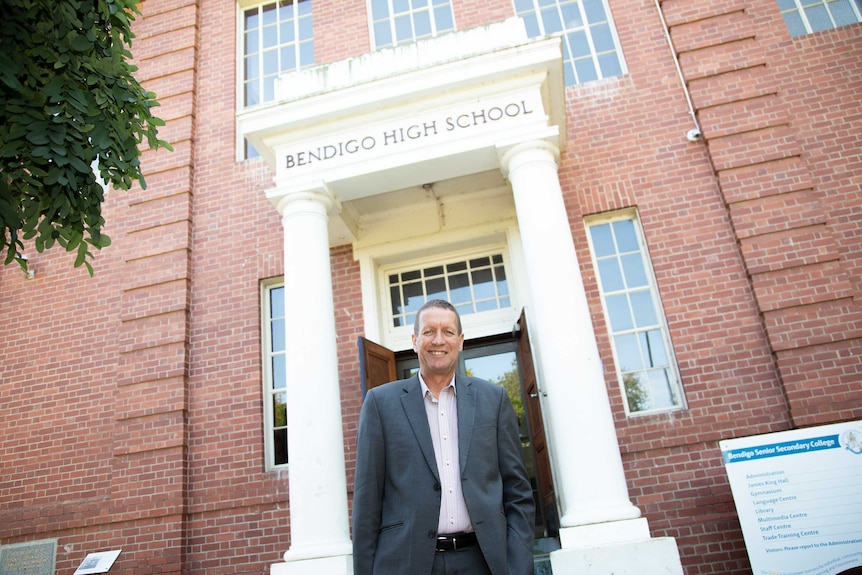 A man with short brown hair stands in front of an old school building.