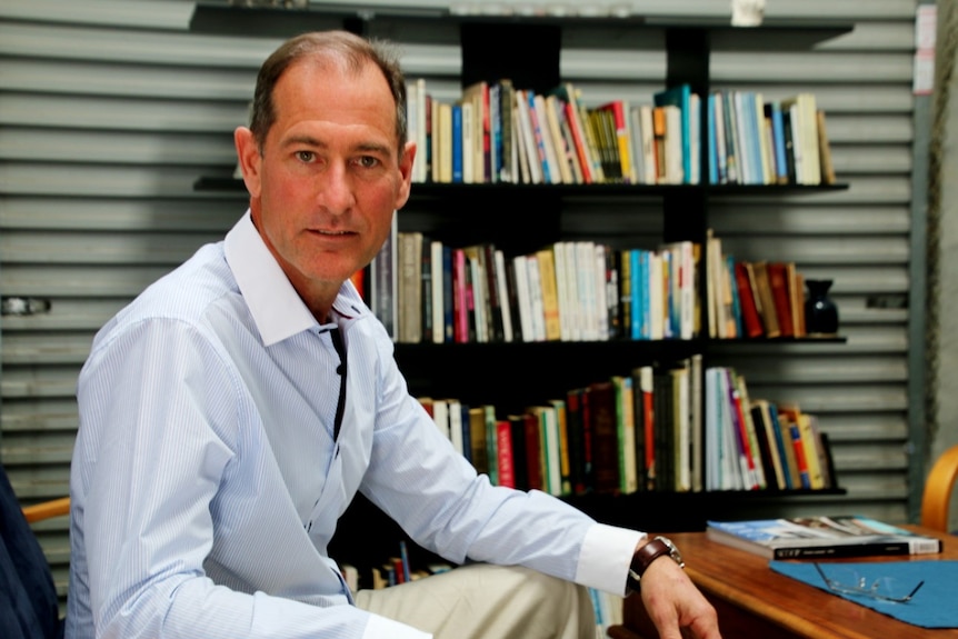 A man wearing an open-necked pale blue collared shirt sits in front of a bookshelf.