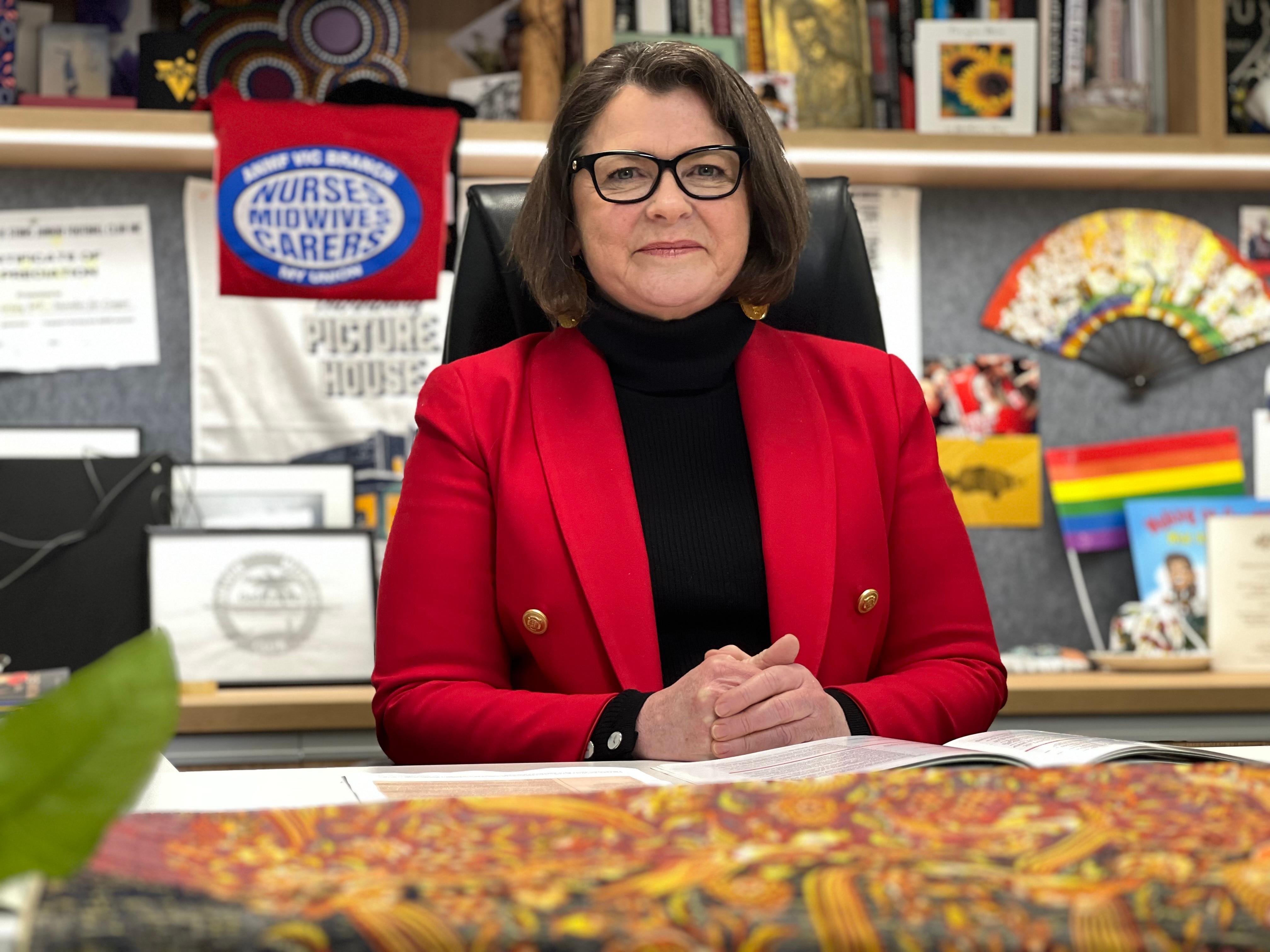Ged Kearney wearing a bright red jacket, sitting at her desk with a nurse union flag in the background.
