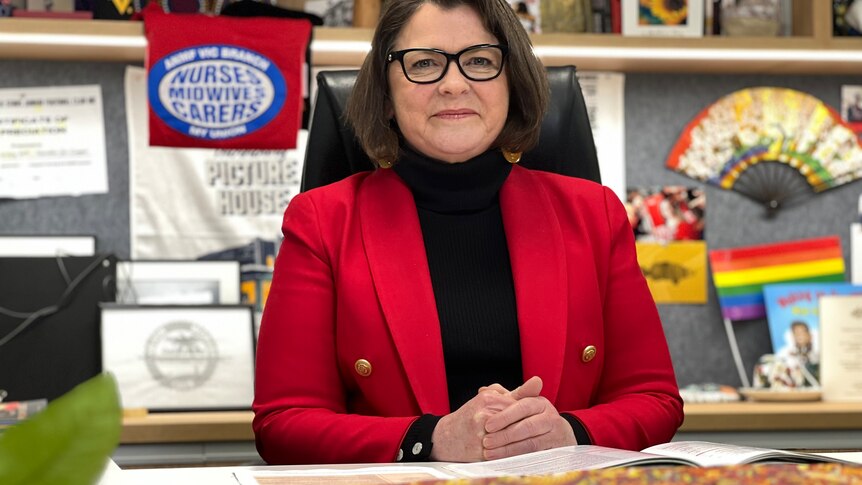 Ged Kearney wearing a bright red jacket, sitting at her desk with a nurse union flag in the background.