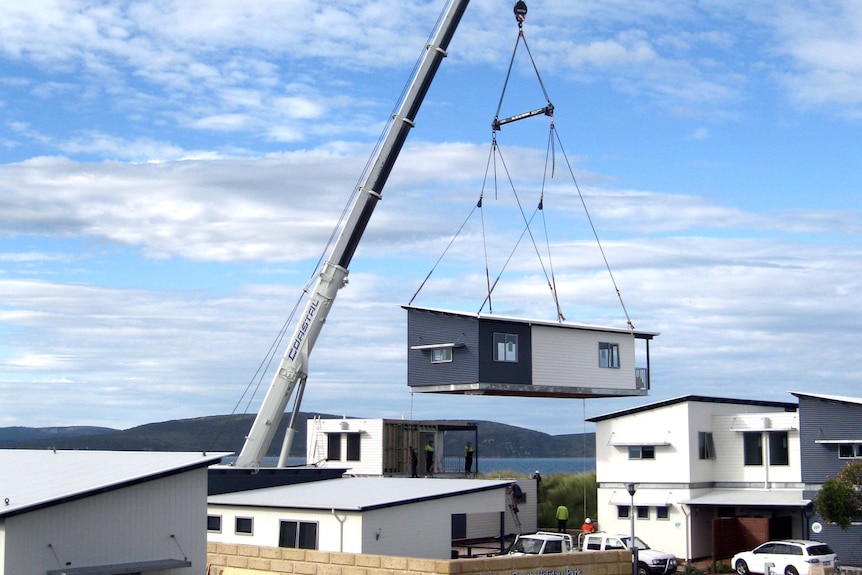 A crane lifting a blue and white modular home between two other similar houses, sea and mountains in the background.