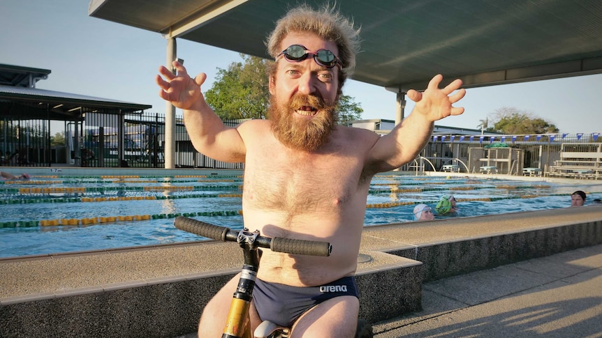 Short-statured man wearing swimming briefs and goggles on his head sits on a tricycle in front of a swimming pool.