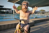 Short-statured man wearing swimming briefs and goggles on his head sits on a tricycle in front of a swimming pool.