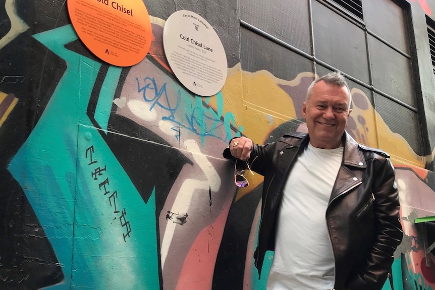 A man in a black leather jacket in front of a mural and a sign on a wall