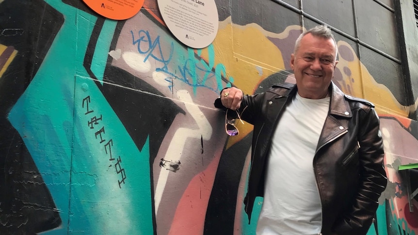 A man in a black leather jacket in front of a mural and a sign on a wall