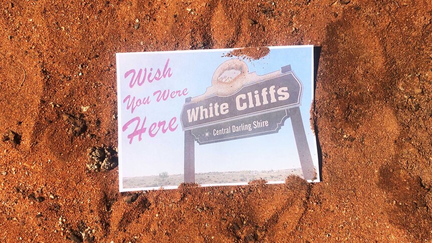 A postcard reading 'wish you were here - White Cliffs" lies in the red dirt.