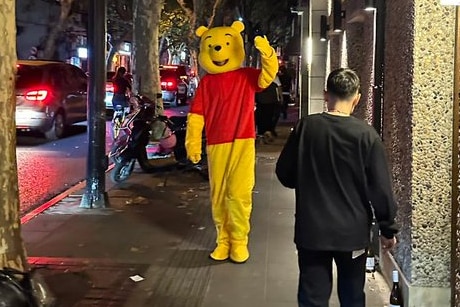 A person wearing a winnie the pooh costume waves as they walk past cars down a busy street at night.
