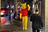 A person wearing a winnie the pooh costume waves as they walk past cars down a busy street at night.