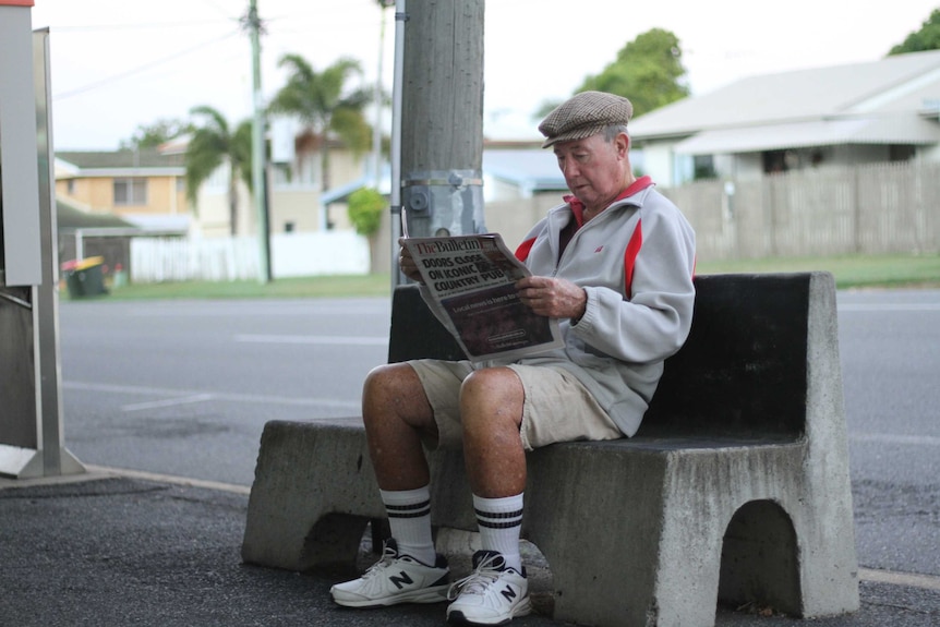 An older man sits on a bench outside a newsagency reading the paper