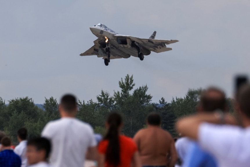 A crowd watches an SU-57 fighter jet fly overhead. 