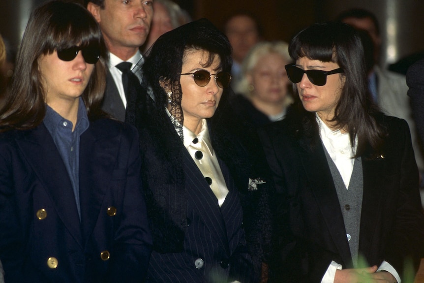 Three women wearing black stand in a funeral crowd. Patrizia Reggiani is wearing a black lace head scarf