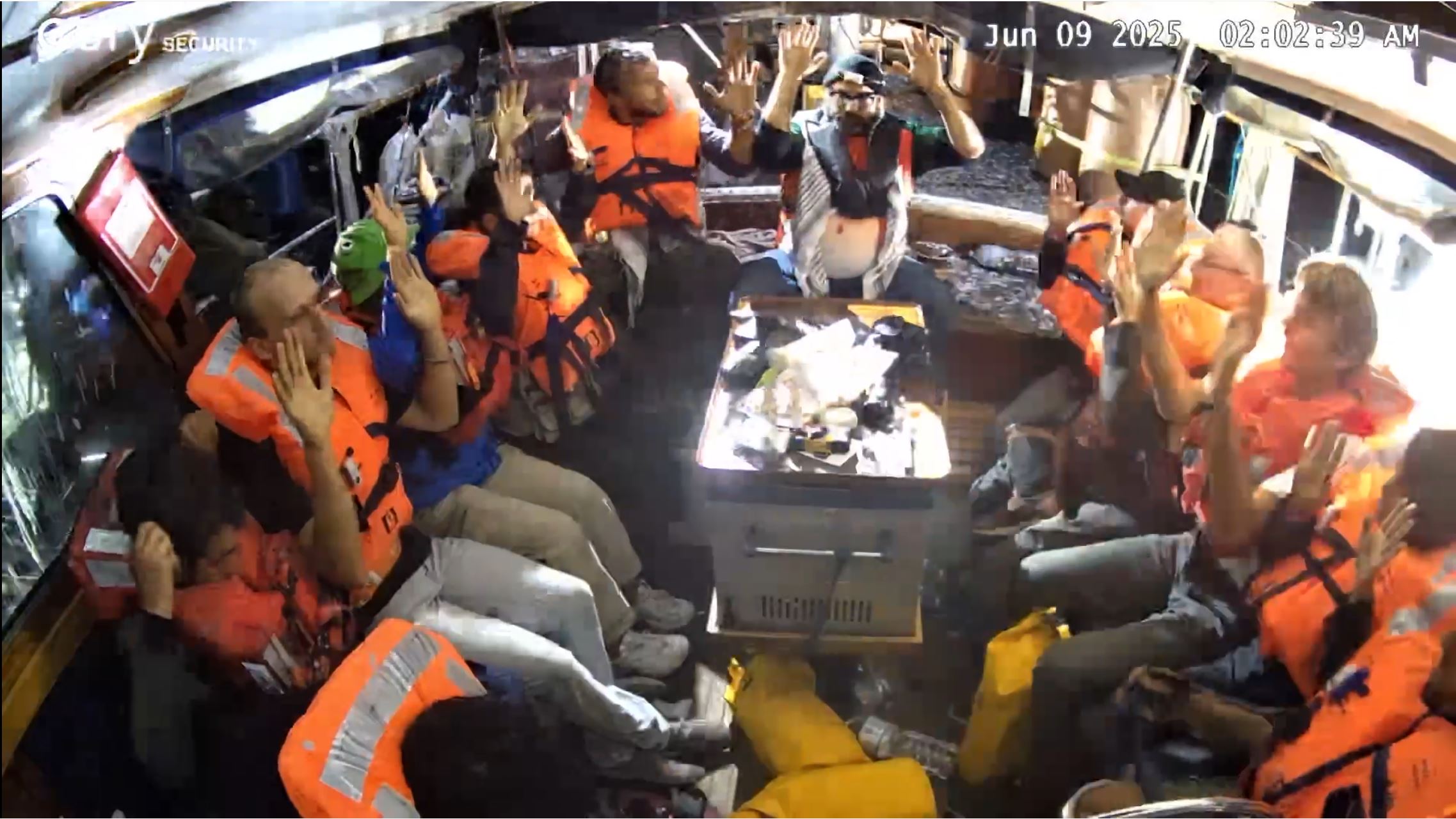 Twelve people wearing orange life jackets sit in a yacht with hands above heads as bright light shines into their ship.