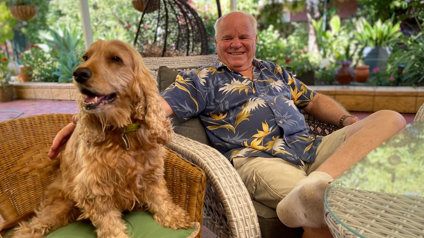 A golden curly haired dog sitting next to a smiling, white balding man wearing a blue, white and yellow button up shirt.