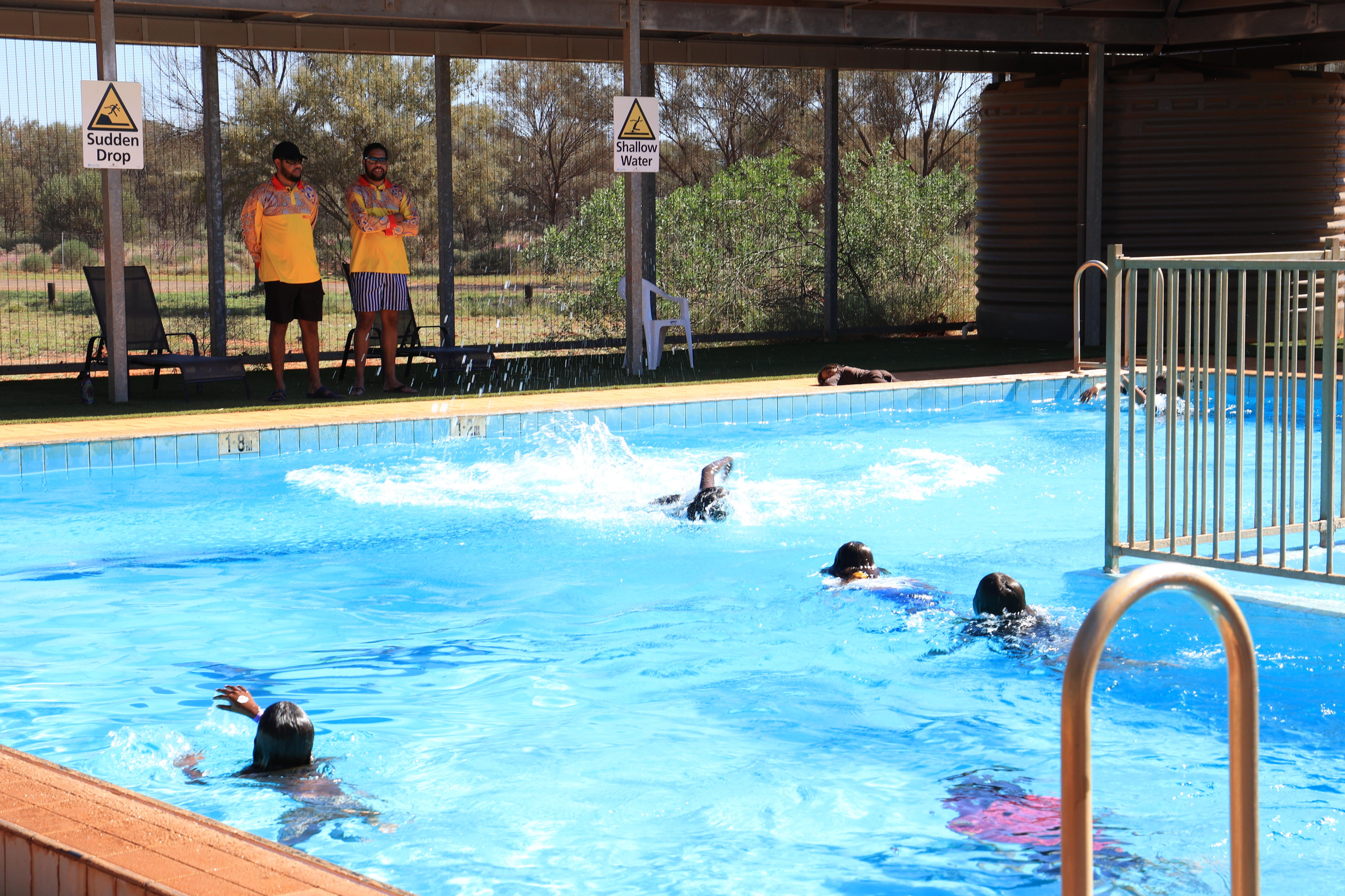 Indigenous children swim in a blue pool, uniformed lifeguards standing under the nearby shade watch on.