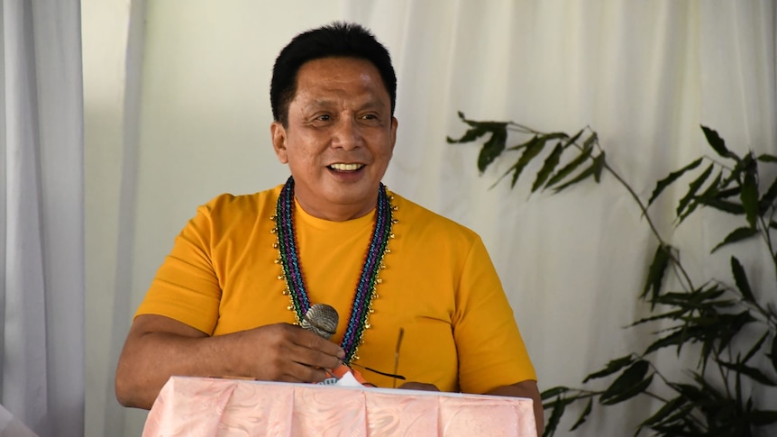  A man wearing an orange top smiles as he holds a microphone.