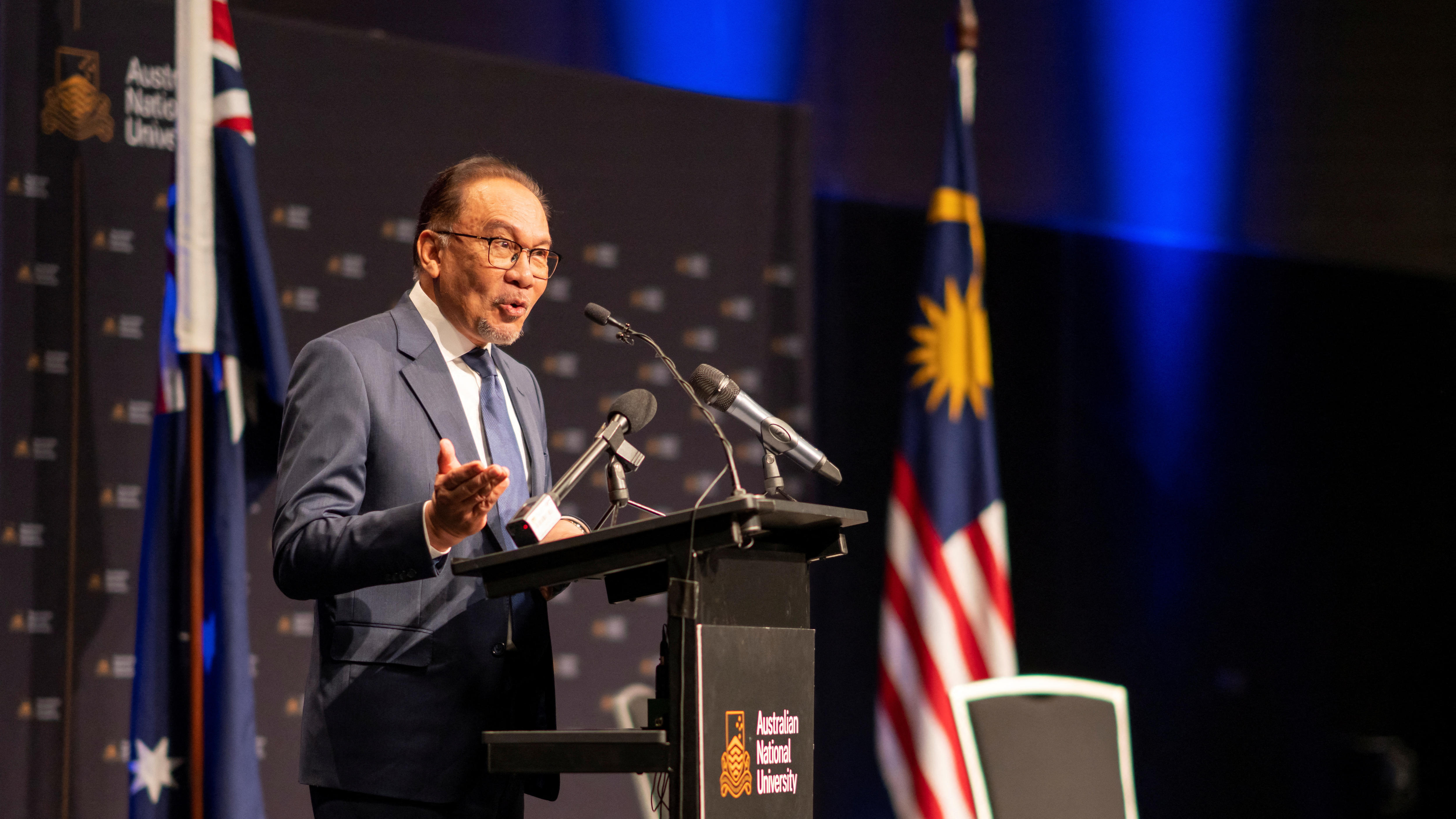 An elderly Asian man in a suit gestures as he gives a talk at a rostrum with flags behind