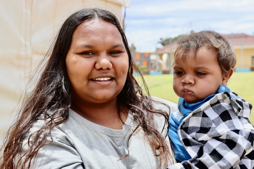 A young Aboriginal mother with long black hair smiles holding her baby boy. 