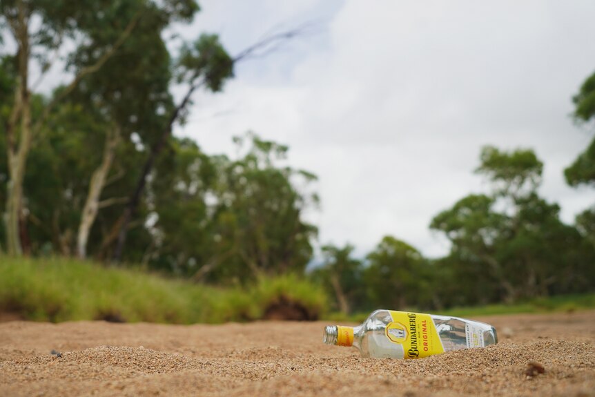 An empty bottle of Bundaberg rum lying on a large patch of sand, surrounded by grass and trees