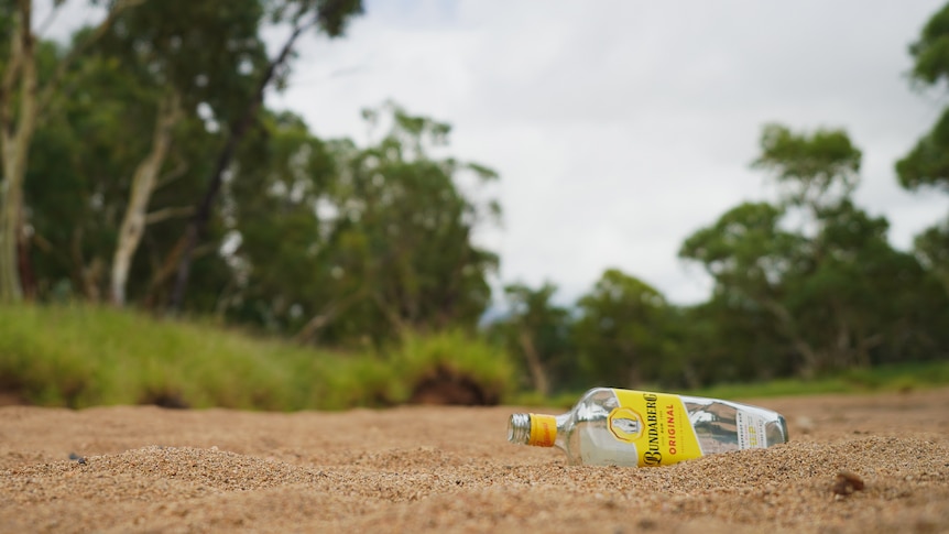 An empty bottle of Bundaberg rum lying on a large patch of sand, surrounded by grass and trees