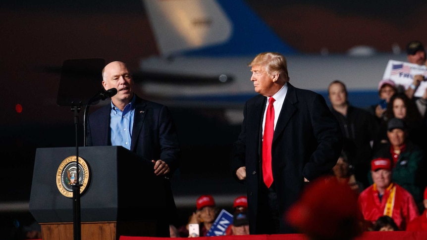 Rep. Greg Gianforte stands in front of a podium next to President Donald Trump.