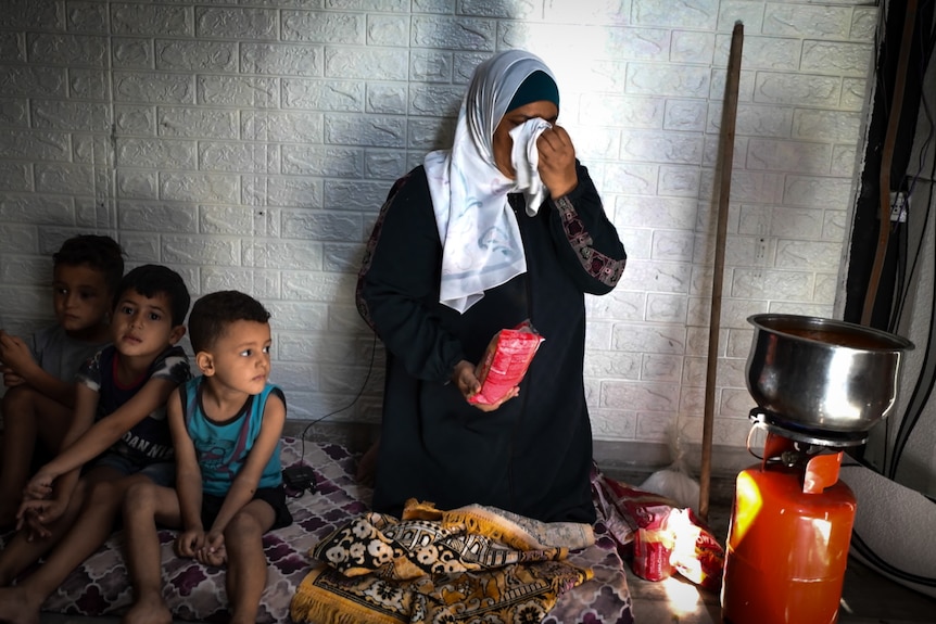 A woman cries into a hanky while three children sit beside her on a blanket. A makeshift stove and gas bottle nearby