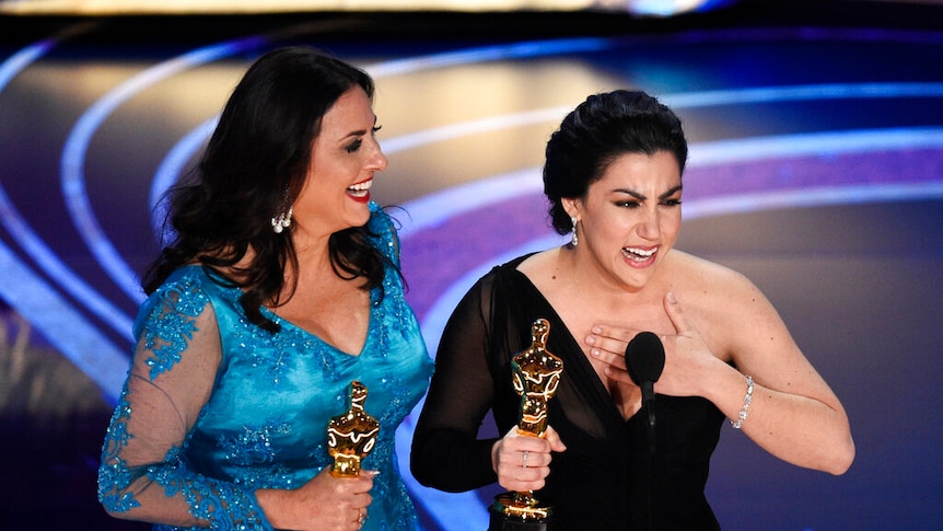 Two women, both wearing floor-length formal gowns, smile while holding Oscar statues.