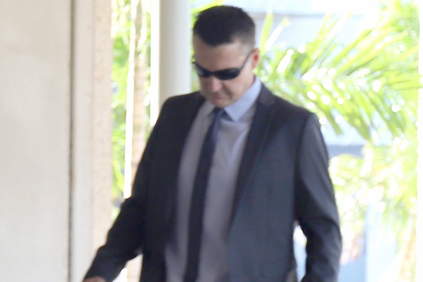 Man in navy suit and white shirt with blue tie and sunglasses walks with head down